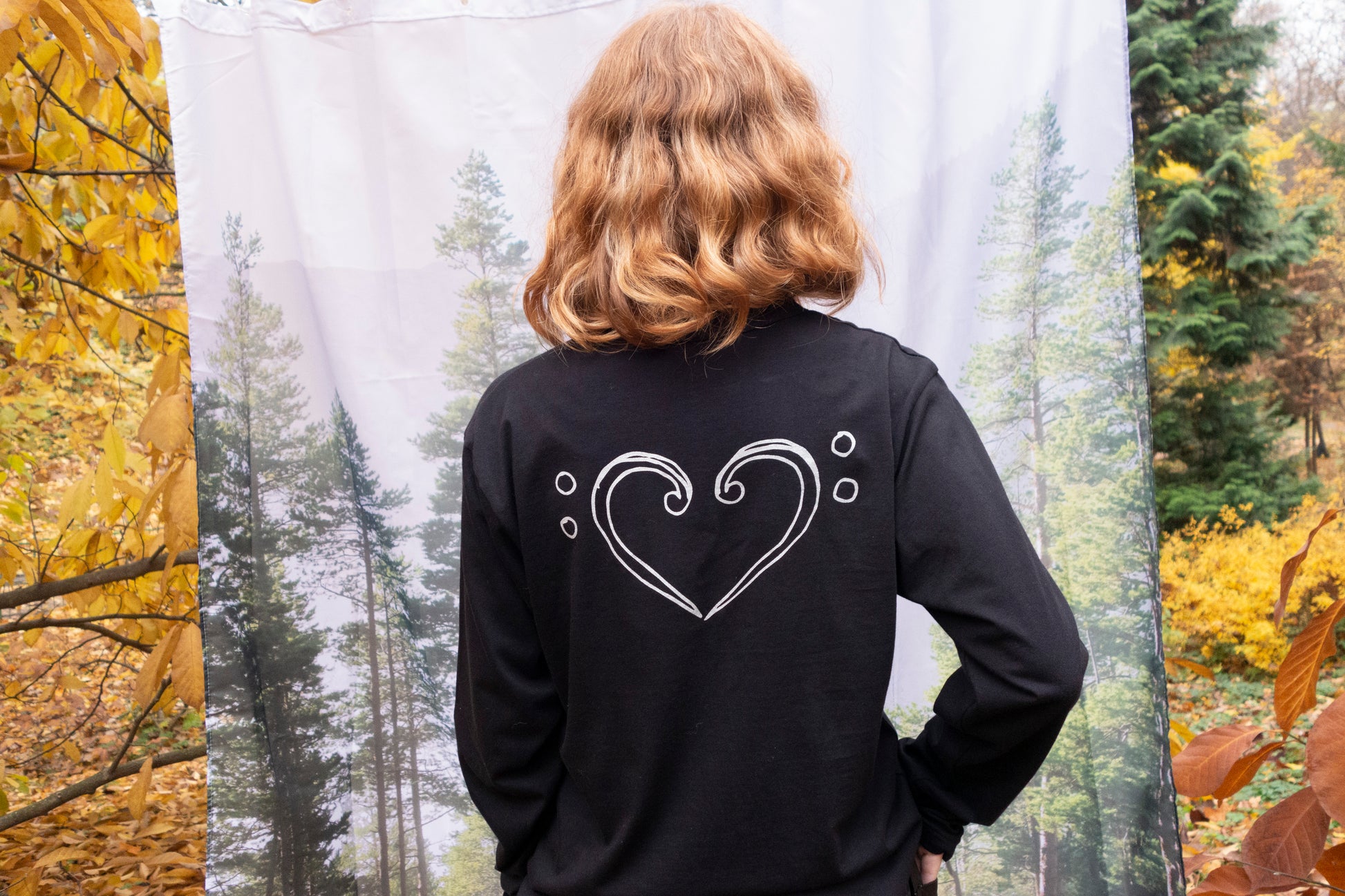 Person wearing a black long-sleeve shirt with a clef heart design in an autumn forest setting