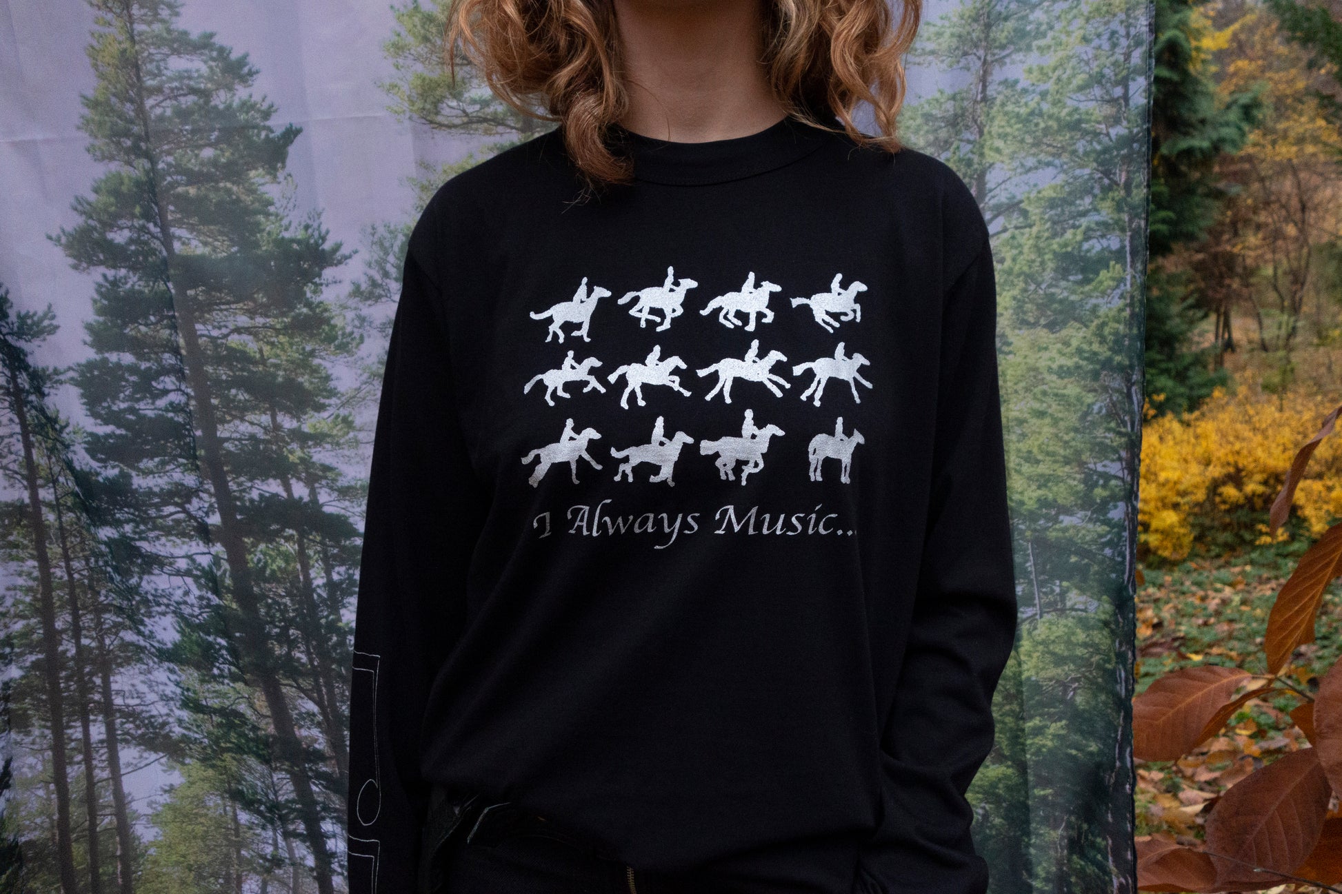 Person wearing a Black long sleeve with silver silkscreen artwork on front, sleeves, and back, designed and produced in Kyiv., standing in front of a forest background.