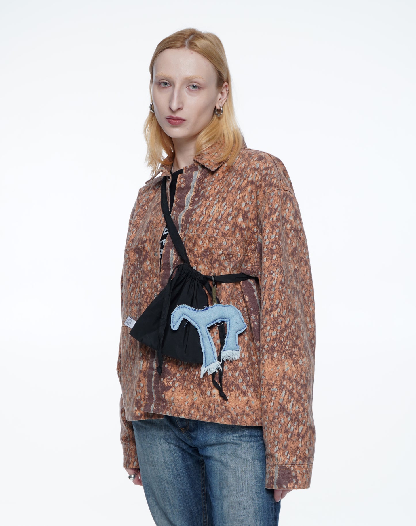 Person wearing a patterned jacket with a black bag featuring a blue horse toy pendant on a white background