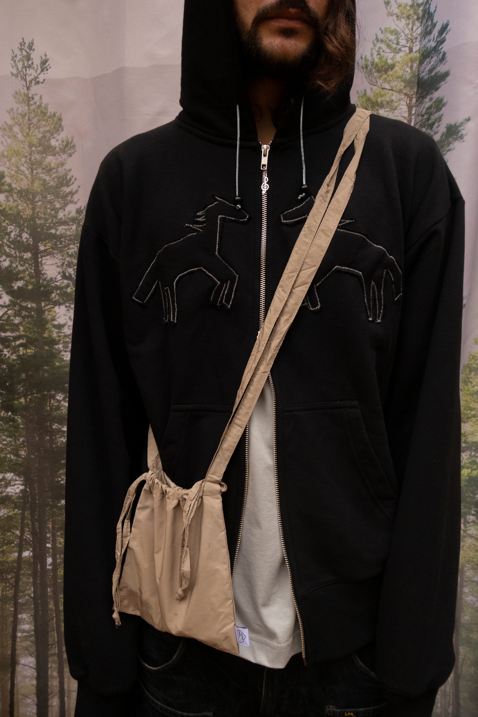 Person wearing a black hoodie with horse graphics and carrying a beige bag in a forest setting