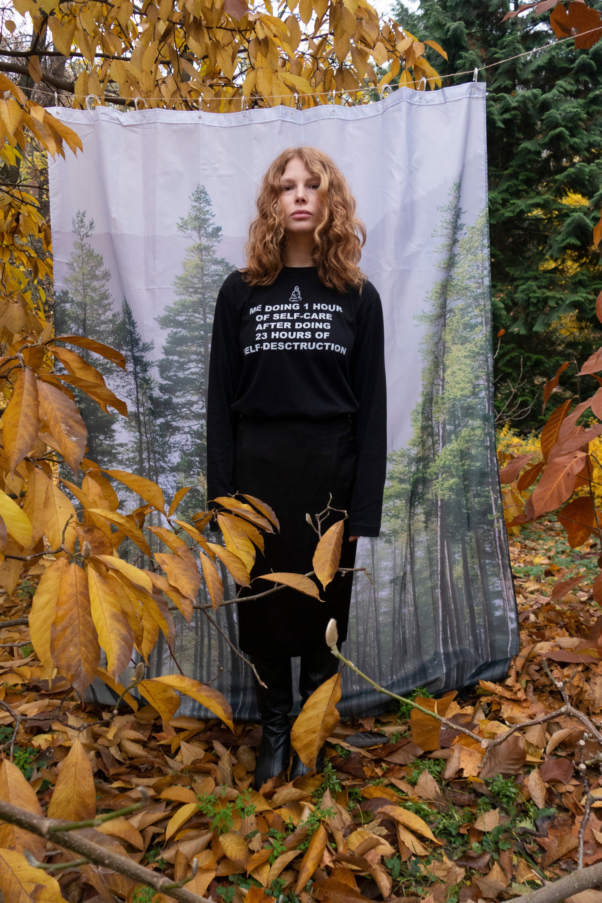 Person wearing a 100% cotton black long sleeve with graphic silkscreen print and text "me doing 1h of selfcare after doing 23h of self-destruction", standing in front of a forest backdrop with autumn leaves.