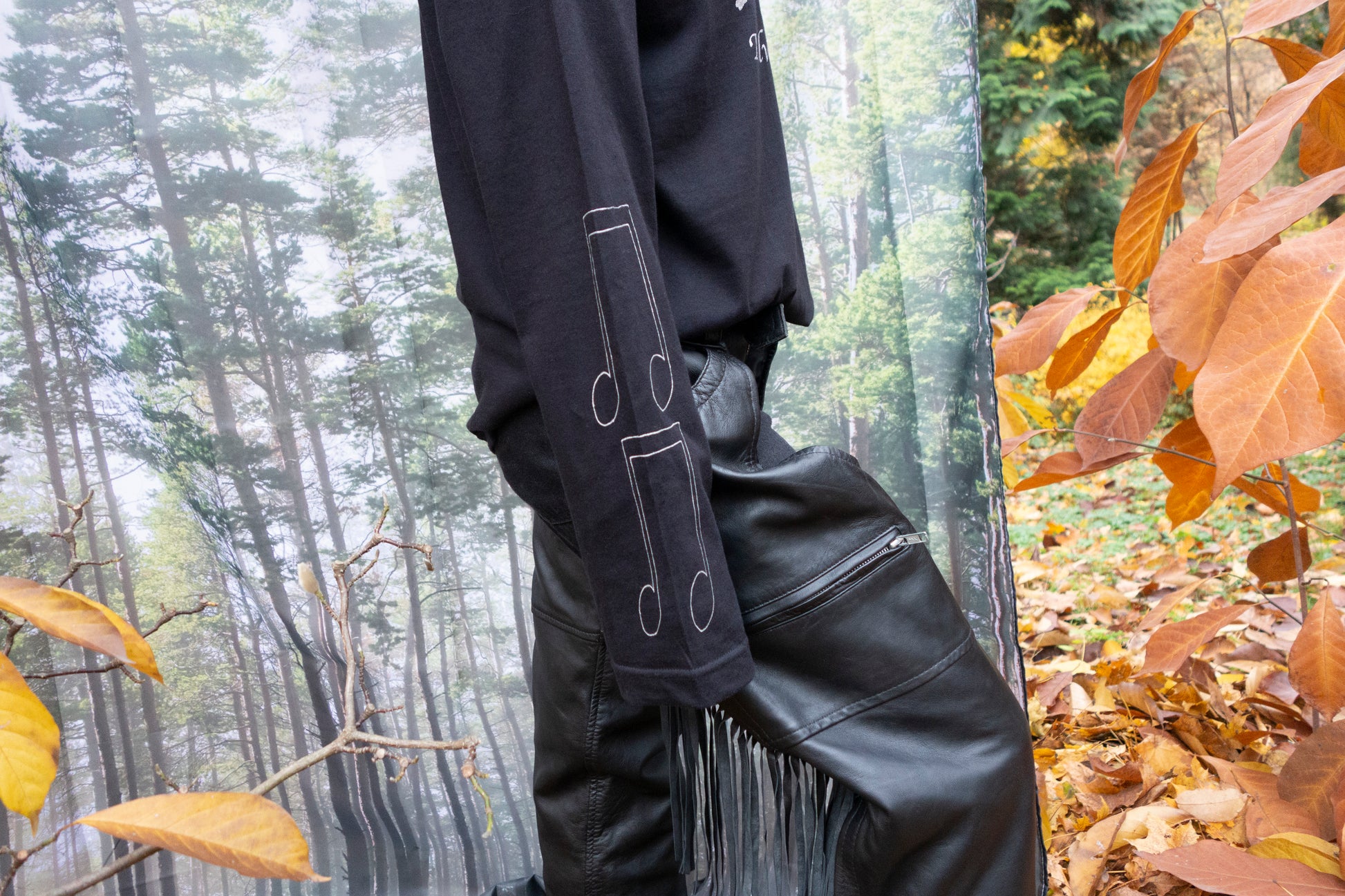 Person wearing black leather pants  with fringe and lack long sleeve T-shirt with silver music-inspired silkscreen prints on front, sleeves, and back in a forest setting